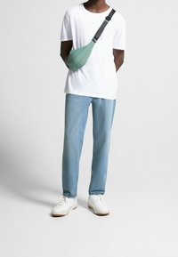 Ljusgrön axelväska av tyg, bärs över kroppen. Modell i en vit t-shirt och ljusblå jeans, står mot en neutral bakgrund.