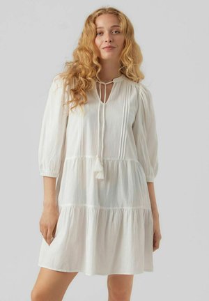 Robe en coton blanc avec un design à étages, présentant un décolleté en cœur, des manches trois-quarts et un lien décoratif avec des franges.