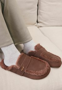 Braune Wildlederslipper mit flauschigem Fellbesatz, einem Riemen über der Oberseite und goldener "SELF"-Markierung, kombiniert mit weißen strukturierten Socken.