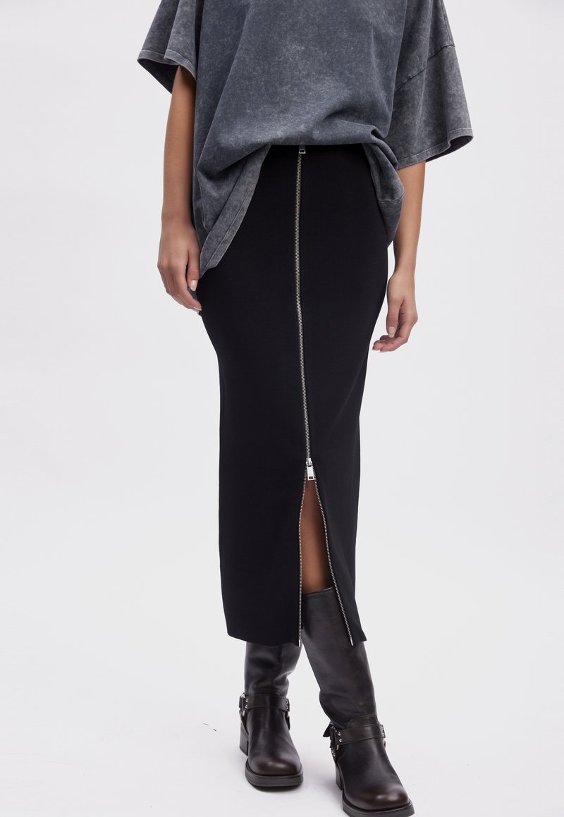 Personne portant une jupe midi noire avec une fente zippée à l'avant, un haut ample gris foncé et des bottes noires mi-mollet à boucle.