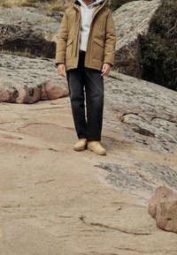 Personne portant une veste couleur fauve, des bottes beiges, un pantalon noir et un sweat à capuche blanc se tient sur un terrain rocheux avec de gros rochers en arrière-plan.