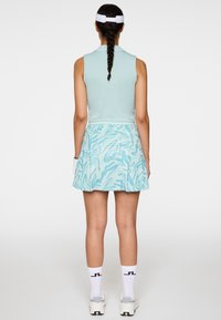 Donna con lunga treccia che indossa una canottiera verde menta, gonna blu con motivo, visiera bianca, calzini e scarpe da ginnastica, in piedi volgendo le spalle.