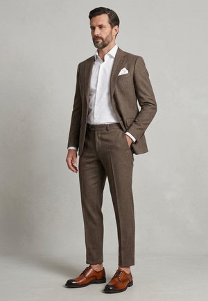 Mann trägt braunen, strukturierten Anzug mit weißem Hemd und Einstecktuch, steht mit einer Hand in der Tasche und braunen Lederschuhen.