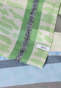 Multikleurrige stof met groene patronen en een grijze strook met pailletten. Heeft een label met de tekst "CECIL". Zachte textuur, gelaagd ontwerp.