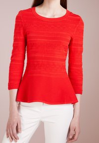 Femme portant un haut péplum rouge texturé à manches longues et un pantalon blanc, debout devant un fond beige.