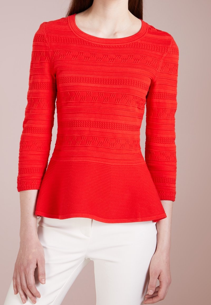 Femme portant un haut péplum rouge texturé à manches longues et un pantalon blanc, debout devant un fond beige.