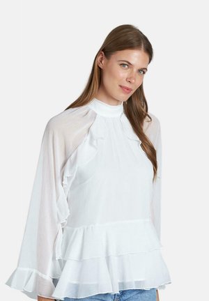 Blusa blanca de peplum con detalles con volantes, cuello alto y mangas transparentes. Textura suave, diseño en capas y ajuste casual con jeans de denim azul.