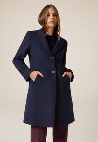 Cappotto in misto lana blu navy con colletto a punta, due tasche frontali e chiusura a tre bottoni. Presenta un tessuto testurizzato e una silhouette aderente.