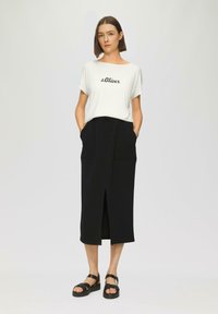 Witte t-shirt met korte mouwen en "s.Oliver" opdruk, gecombineerd met een zwarte rokken tot de knie met een vooropening en zakken. Draagt zwarte sandalen.