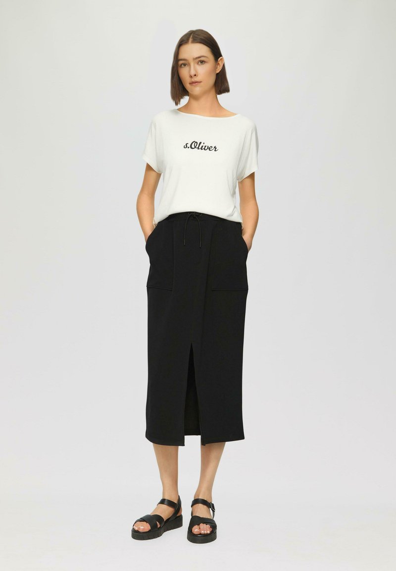 Witte t-shirt met korte mouwen en "s.Oliver" opdruk, gecombineerd met een zwarte rokken tot de knie met een vooropening en zakken. Draagt zwarte sandalen.