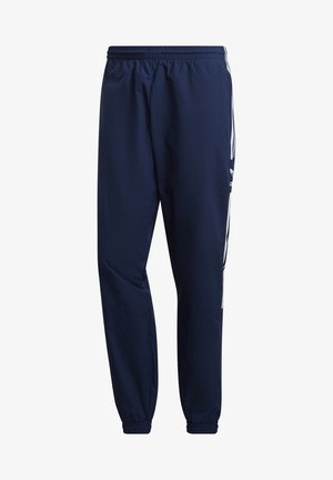 Donkerblauwe trainingspak van lichtgewicht stof met witte drie-strepen accenten op de mouwen van de jas en de broek, gecombineerd met klassieke witte sneakers.