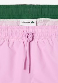 Rosa svømmedrakt med elastisk liv, grønn innside og snøring. Har Lacoste-logo. Glatt, lett stoff.