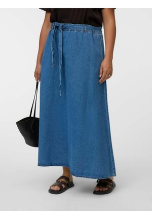 Person trägt ein schwarzes Oberteil und einen langen blauen Jeansrock mit Bindegürtel, hält eine schwarze Tasche, trägt schwarze Sandalen und steht vor einem schlichten Hintergrund.
