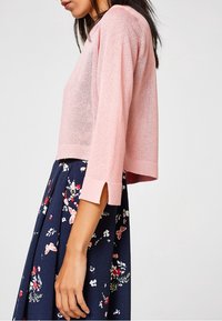 Světlounce růžový krátký svetr s tříčtvrtečními rukávy z texturovaného materiálu, kombinovaný s květinovou tmavě modrou sukní s výraznými vzory.
