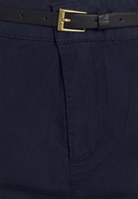 Námořnické chinos s hladkým povrchem, s černým koženým opaskem se zlatým přezkou a jedním detailem přední kapsy.