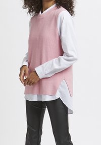 Maglione rosa senza maniche sopra una camicia bianca, caratterizzato da spacchi laterali per facilitare i movimenti e un collo rotondo. Pantaloni di pelle nera.