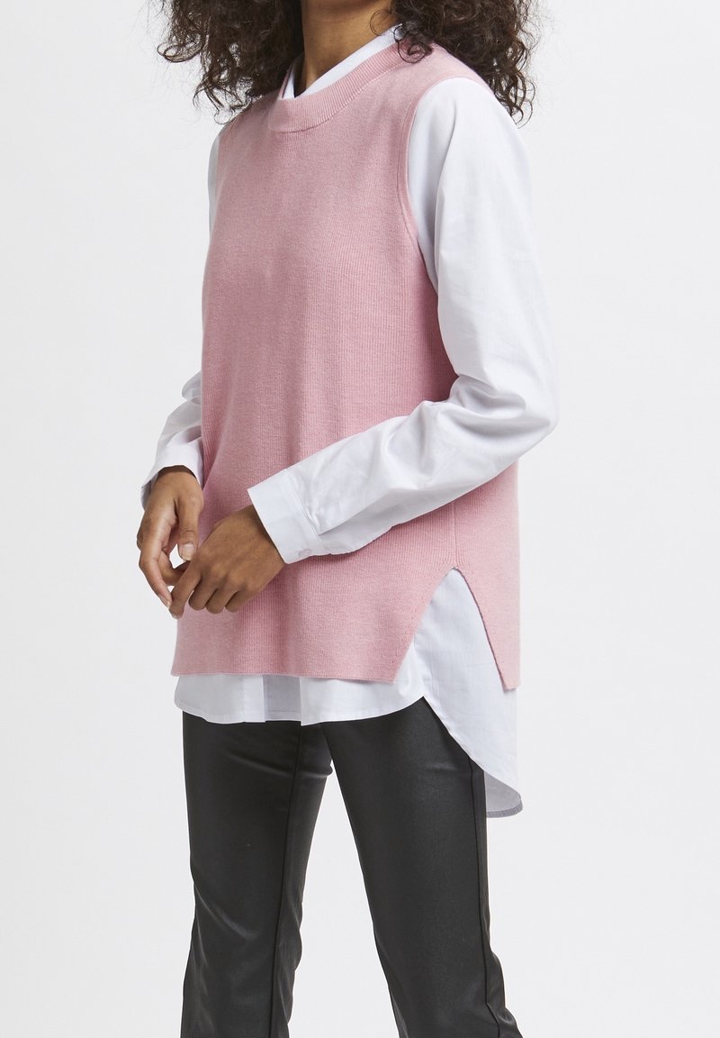 Maglione rosa senza maniche sopra una camicia bianca, caratterizzato da spacchi laterali per facilitare i movimenti e un collo rotondo. Pantaloni di pelle nera.