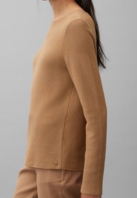 Sweter dzianinowy w jasnobrązowym kolorze, z teksturowaną powierzchnią, długimi rękawami i okrągłym dekoltem. Subtelny detal w postaci guzika u dołu.