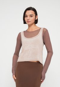 Top court beige clair sans manches avec un col arrondi, présentant un motif texturé et une sous-couche marron à manches. Porté avec une jupe marron.