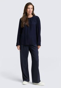 Marineblaues Langarmshirt mit gerafftem Ausschnitt, kombiniert mit passenden weit geschnittenen Hosen. Glatter Stoff und entspannte Passform, ergänzt durch weiße Schuhe.