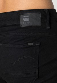 Jean en denim noir avec une finition texturée, doté d'une pièce en cuir marquée "RAW" à la ceinture et d'une petite étiquette sur une poche arrière.