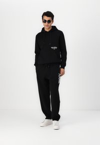 Čierna mikina s kapucňou a detailom loga, v kombinácii s čiernymi teplákmi, ktoré majú biely text. Biele tenisky dopĺňajú outfit.