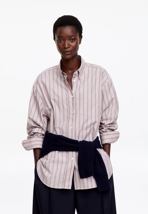 Vrouw met kort haar die een losse gestreepte blouse met knopen draagt met opgerolde mouwen en een donker vest geknoopt rond haar taille.