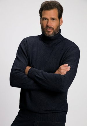 Maglione a dolcevita blu scuro in maglia strutturata con polsini e colletto a costine. Il modello posa con le braccia incrociate su uno sfondo neutro.