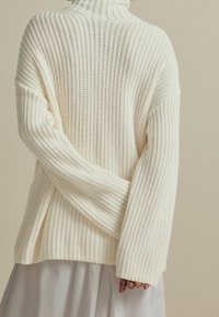 Person in einem cremefarbenen, grob gestrickten Pullover mit langen Ärmeln und einem weißen Rock, die Arme hinter dem Rücken verschränkt.
