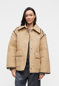 Μπεζ μπουφάν με καπιτονέ, γιακά, με υφασμάτινο μοτίβο και μαύρες διακοσμητικές λεπτομέρειες, δύο τσέπες στο μπροστινό μέρος και χαλαρή εφαρμογή.