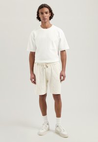 Tricou alb din bumbac cu mâneci scurte și un buzunar pe piept, purtat cu pantaloni scurți bej deschis, captușați, cu talie ajustabilă. Tenisi albi.