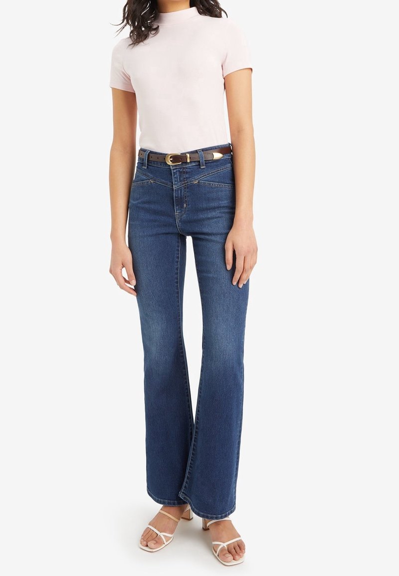 Mujer con top de manga corta color rosa claro, jeans de talle alto azul con un cinturón marrón oscuro y sandalias de tacón blancas con tiras.