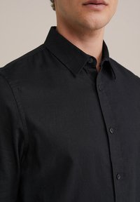 Črna srajca s kratkimi rokavi in ovratnikom, izdelana iz gladkega materiala. Ima sprednje gumbe in vidne šive na ovratniku in rokavih.