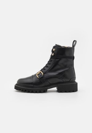 Botte de combat en cuir noir avec semelle à crampons, sangle à boucle à la cheville, fermeture à lacets sur le devant, œillets métalliques et languette de tirage sur fond uni.