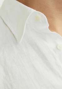Weißes Leinenhemd mit einem Button-Down-Kragen, das eine weiche Textur aufweist und eine Nahaufnahme zeigt, die die Kragen- und Knopfdetails hervorhebt.