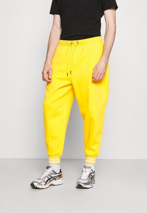 Gele joggingbroek met een elastische tailleband en zijzakken; textiel met een stevige structuur en taps toelopende boorden, gecombineerd met grijze sportschoenen.
