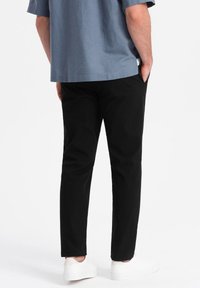 Pantalons noirs ajustés, en tissu lisse. Comprend des poches latérales et portés avec des baskets blanches. Vue arrière montrant un design simple.