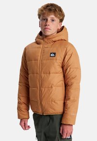 Brązowa pikowana kurtka puchowa z kapturem, z zamkiem błyskawicznym z przodu, pikowanym wzorem i naszywką z logo na piersi. Nosi się ją na zielonych spodniach.