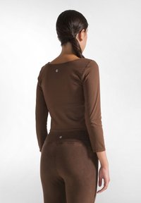 Maglione marrone a maniche lunghe con scollatura ampia, texture liscia e dettaglio con logo sul retro, abbinato a pantaloni attillati coordinati.