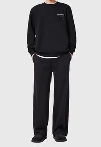 Persona con sudadera negra de AllSaints, pantalones anchos negros, zapatos negros y una camiseta blanca visible en el dobladillo, de pie con las manos en los bolsillos.
