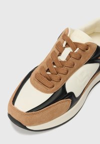 Il design delle sneaker presenta una tomaia in tessuto bianco con accenti in suede marrone e elementi in pelle nera, completato da lacci marroni e una sobria brandizzazione.