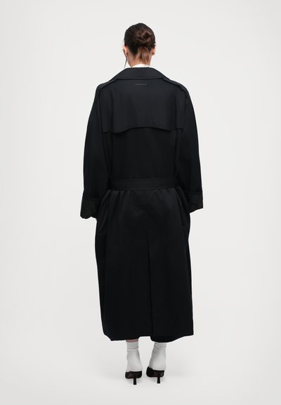 Lång svart trenchcoat med bred krage, knytmidja och sidofickor. Har en plisserad rygg och långa ärmar med uppvikt manschett.