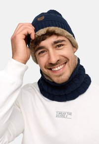 Navy knit beanie with tan fleece lining and matching neck gaiter, featuring ribbed texture and a decorative logo patch. White long-sleeve shirt visible.