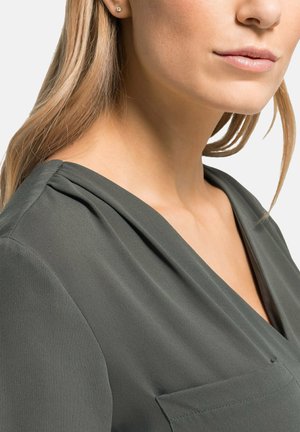 Dunkelgrüne Bluse mit V-Ausschnitt, aus weichem, fließendem Stoff und Raffung an den Schultern. Eine Tasche ist an der Vorderseite sichtbar.