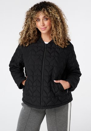 Vrouw met krullend haar draagt een zwarte gewatteerde jas met hartpatronen, handen in de zakken, en grijze broek met witte zijstrepen.