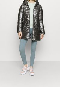 Lesklý černý prošívaný kabát s kapucí, zapínáním na zip a dvěma předními kapsami, nošený přes světle zelené tričko a modré legíny s pastelovými teniskami.