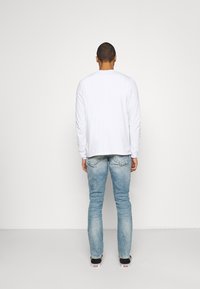 Vit långärmad skjorta med rund halsringning, matchad med ljusblå jeans och svarta sneakers, sedd bakifrån mot en enfärgad bakgrund.