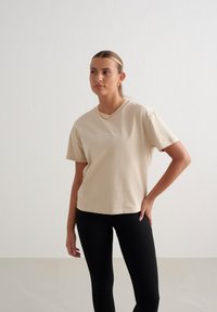 Beige Baumwoll-T-Shirt mit kurzen Ärmeln, klassischem Rundhalsausschnitt und dezentem weißen Logotext. Kombiniert mit schwarzen Ripp-Leggings.