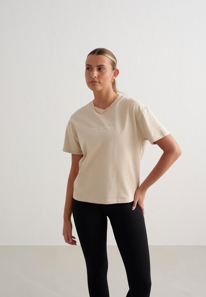 Beige Baumwoll-T-Shirt mit kurzen Ärmeln, klassischem Rundhalsausschnitt und dezentem weißen Logotext. Kombiniert mit schwarzen Ripp-Leggings.