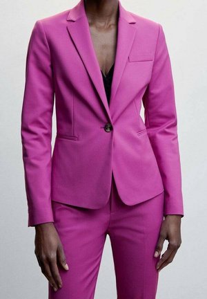 Kvinde iført en tætsiddende magenta blazer og matchende bukser med en sort top nedenunder, stående mod en ensfarvet lys baggrund.